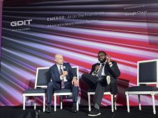 David Miller, interim chief technology officer at the FBI, is pictured sitting in a chair on stage at a GDIT event. He is holding a microphone and sits in front of a striped backdrop with the name of the event on it: "EMERGE: AI on the Frontlines."